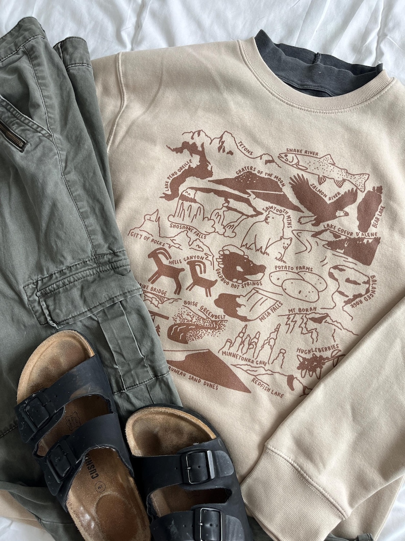 May include: A tan sweatshirt with a brown graphic print of a map of Idaho. The map includes the names of various landmarks and natural features, such as the Snake River, Shoshone Falls, and the Tetons. The sweatshirt is paired with a pair of green cargo pants and black sandals.