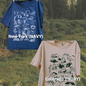 May include: Two t-shirts with graphic designs of mountain ranges and landmarks. The shirt on the left is navy blue with white graphics and says "New York (NAVY)". The shirt on the right is ivory with green graphics and says "Colorado (IVORY)".