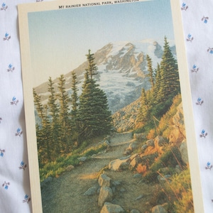May include: A vintage postcard featuring a scenic view of Mount Rainier National Park, Washington. The image showcases a hiking trail leading towards a snow-capped mountain, surrounded by evergreen trees and rocky terrain. The postcard has a cream-colored border.