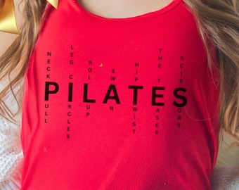 May include: Red tank top with a black word search puzzle that spells out "PILATES" in the center. The puzzle includes words related to pilates exercises, such as "neck", "leg", "roll", "hip", "waist", and "scissors".