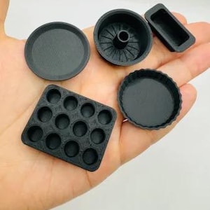 Könnte beinhalten: Eine Sammlung kleiner, schwarz 3D-gedruckter Backformen und -tabletts. Das Set enthält ein rundes Tablett, eine Tarteform, eine rechteckige Form, eine Muffinform und eine Gugelhupfform, alles in einer Hand gehalten.