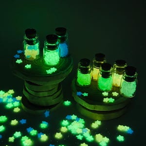 May include: A collection of small, clear glass bottles with dark cork stoppers, each filled with tiny, glowing star-shaped ornaments. The stars emit a vibrant green and blue light, and many loose glowing stars are scattered around the base.