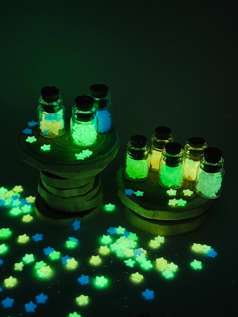 May include: A collection of small, clear glass bottles with dark cork stoppers, each filled with tiny, glowing star-shaped ornaments. The stars emit a vibrant green and blue light, and many loose glowing stars are scattered around the base.