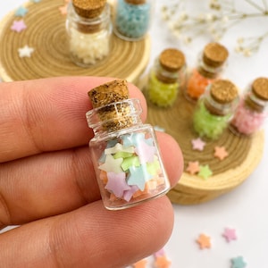 May include: A miniature clear glass bottle with a cork stopper filled with tiny pastel-colored star-shaped beads in shades of pink, blue, yellow, and green. Several other identical glass bottles filled with stars are arranged on wooden discs.