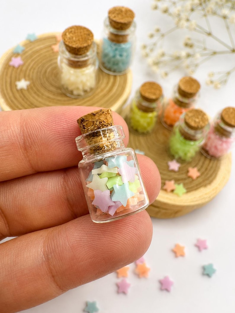 May include: A miniature clear glass bottle with a cork stopper filled with tiny pastel-colored star-shaped beads in shades of pink, blue, yellow, and green. Several other identical glass bottles filled with stars are arranged on wooden discs.