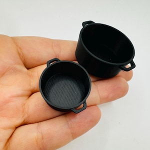 Könnte beinhalten: Zwei kleine, schwarze, runde Töpfe mit Griffen. Ein Topf hat eine gerippte Innenseite, der andere ist glatt. Die Töpfe sind wahrscheinlich Miniatur- oder Spielzeugkochgeschirr.