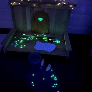 May include: A wooden miniature house structure featuring an arched door with a glowing green heart cutout. The scene includes scattered glowing green star-shaped accents, a white bone-shaped sign, a small dark container, and two white boots.