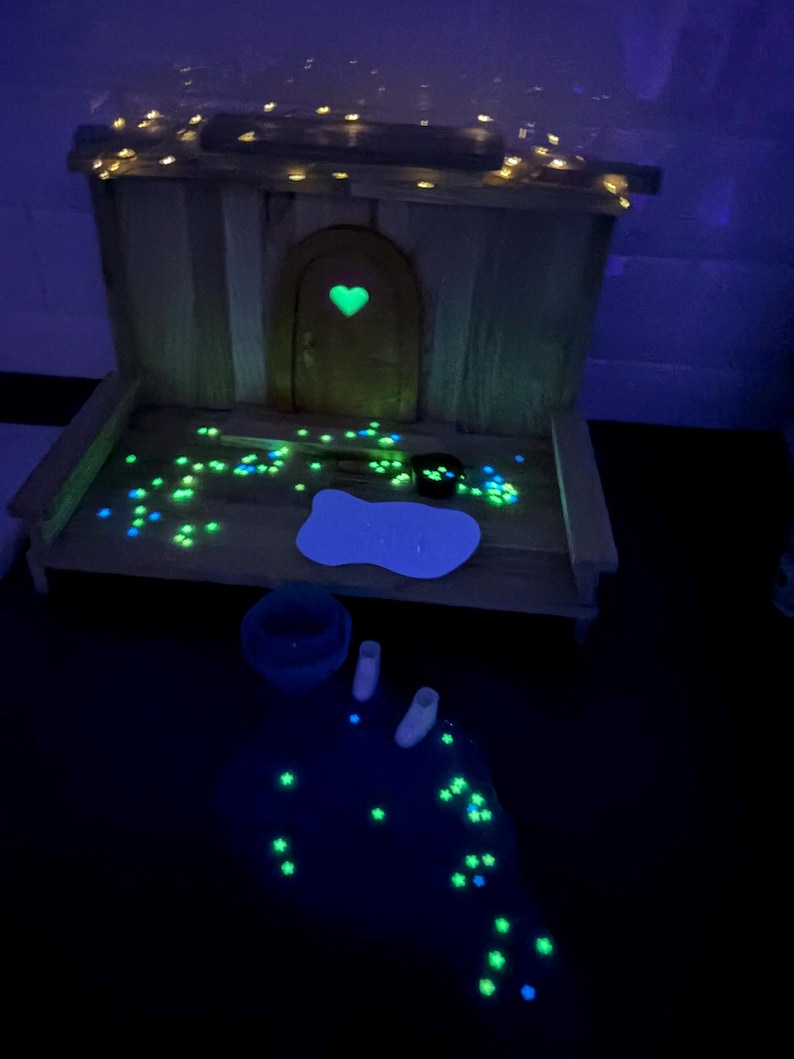 May include: A wooden miniature house structure featuring an arched door with a glowing green heart cutout. The scene includes scattered glowing green star-shaped accents, a white bone-shaped sign, a small dark container, and two white boots.