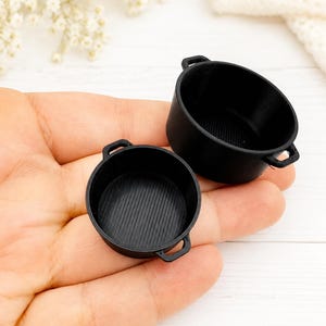 Könnte beinhalten: Zwei schwarze Miniatur-Kochtöpfe mit runden Körpern und kleinen Seitengriffen. Jeder Topf verfügt über einen texturierten, gerippten Innenboden und eine glatte Außenfläche. Diese kleinen, oben offenen Gefäße haben eine kreisförmige Form.