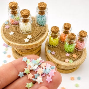 May include: Small clear glass bottles with cork stoppers filled with tiny pastel-colored star-shaped decorations. The stars are in shades of pink, blue, yellow, green, and white. Some stars are scattered outside the bottles on the surface.