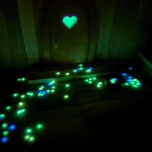 May include: A miniature arched door featuring a glowing green heart-shaped cutout at the center. The ground in front of the door is scattered with numerous small, luminous star-shaped accents in shades of bright green and blue, creating a trail.