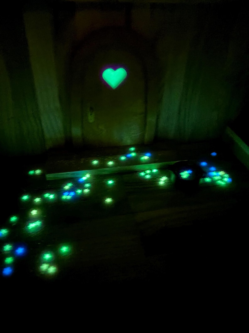 May include: A miniature arched door featuring a glowing green heart-shaped cutout at the center. The ground in front of the door is scattered with numerous small, luminous star-shaped accents in shades of bright green and blue, creating a trail.