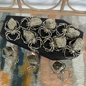 May include: A collection of silver spider figurines with pyrite stones for their bodies. The spiders are arranged on a black velvet coffin-shaped box.