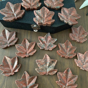 May include: A set of 13 red and grey maple leaf shaped coasters made of polished stone. The coasters are arranged on a wooden surface.