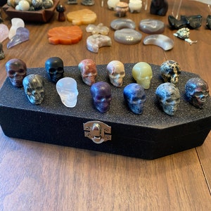 May include: A black velvet coffin-shaped box with a silver latch, filled with 13 small, polished skull-shaped stones in various colors, including clear, purple, green, brown, and black.