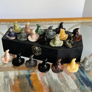 May include: A collection of 18 small, hand-carved witch hats made from various types of stone. The hats are displayed in a black velvet box with a silver latch. The hats are in a variety of colours, including green, brown, black, white, and red.