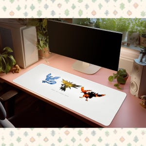 May include: A white rectangular desk mat featuring three stylised bird illustrations in blue, yellow, and orange-red, arranged horizontally. The design includes small text below the figures that reads "WHERE THE LEGENDS BEGIN".