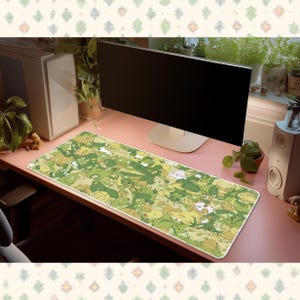 May include: A rectangular desk mat featuring a dense, repeating pattern of various green and yellow cartoon creatures and plant-like characters. The mat has a smooth surface with a white border and is positioned on a flat, solid pink surface.
