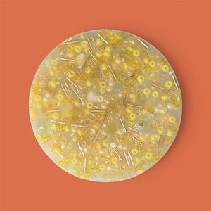 May include: A round, light green object filled with yellow and gold beads. The beads are various sizes and shapes, including small spheres and elongated cylinders. The background is a solid orange color.