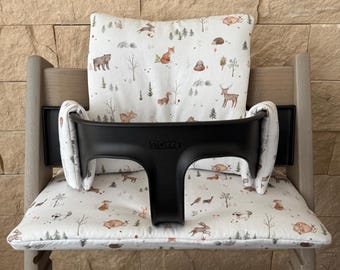 May include: A wooden high chair with a black safety bar and forest-themed cushions. The cushions feature a white background with illustrations of woodland animals, including bears, foxes, and deer. The chair is designed for a baby or toddler.