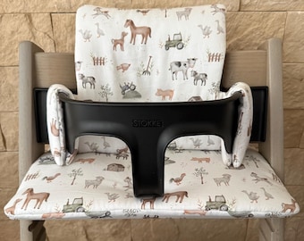 May include: A wooden high chair with a black safety bar and a farm animal-themed cushion set. The cushion features illustrations of horses, pigs, tractors, and other farm elements. The black safety bar has the brand name "STOKKE" printed on it.