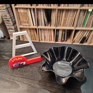 May include: A black, upcycled vinyl record bowl with a wavy edge sits on a dark surface. A red and black Little Shop of Horrors themed item and a white letter A are also present. Shelves of vinyl records are in the background.