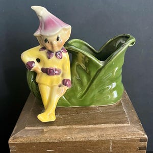 May include: A vintage ceramic figurine of a whimsical elf in a yellow outfit with purple accents, leaning against a green leaf-shaped planter. The elf has a pink hat and a cheerful expression. The planter is a vibrant green color.