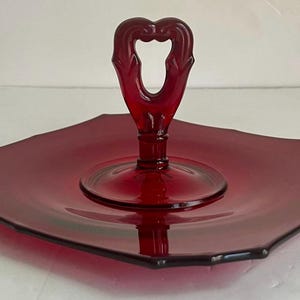 Puede incluir: Bandeja de servir de cristal rojo rubí con asa en forma de corazón. La bandeja tiene forma cuadrada con bordes festoneados y base circular. El asa es ornamentada. Una pieza vintage.