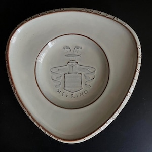Può includere: Piatto triangolare in ceramica color crema con bordo marrone e la scritta "HERRING" sul bordo. Il centro presenta un emblema dettagliato con la scritta "HEERING" sottostante. Il piatto ha una superficie leggermente irregolare.