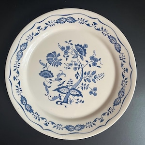Puede incluir: Un plato de cerámica blanco con un diseño floral azul. El plato presenta un motivo floral central y un borde decorativo con hojas y flores. El diseño es de estilo clásico y vintage.