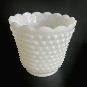 May include: A white hobnail vase with a scalloped rim. The vase has a textured surface with a pattern of raised dots and horizontal lines. The vase is opaque and sits on a black surface.