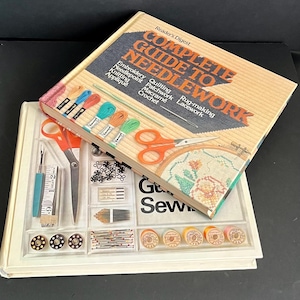 Op de afbeelding: Twee boeken, waarvan één met de titel "Reader's Digest Complete Guide to Needlework", met kleurrijk borduurgaren en scharen. Het andere boek bevat naaibenodigdheden zoals spoelen, naalden en een meetlint. De boeken zijn beige en oranje.