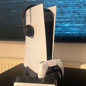 Könnte beinhalten: Eine weiße PlayStation 5 Konsole mit einem schwarzen Headset und einem weißen Controller auf einem schwarzen Ständer. Der Ständer trägt den Text "DARKMATTERY1900".
