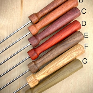May include: Seven fondue forks with wooden handles in various colors, including dark brown, reddish-brown, and light tan. The metal prongs are silver. The handles are labeled A through G.