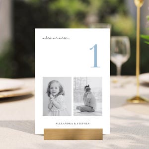 May include: A white table card with the text "when we were..." and the number "1" in light blue. Two black and white photos are below the number. The names "ALEXANDRA & STEPHEN" are at the bottom. The card is in a wooden holder.