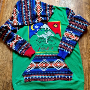 May include: Green hoodie with patterned sleeves and hood. The front features a graphic of a person riding a bicycle with a mountain, sun, and moon. The year "1943" is printed below the graphic. The sleeves and hood have a blue, red, and white geometric design.
