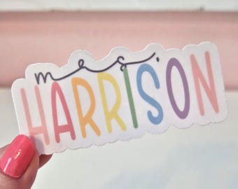 May include: A colorful sticker with the name "Harrison" in rainbow hues. The word is written in a playful font, with a small "m" above the "H". A black line with decorative curves sits above the name. The sticker is held by a person with pink nail polish.