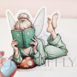 May include: A sticker featuring a fairy reading a book. The fairy is wearing a green outfit and has blonde hair in a bun. She is lying on a mushroom-shaped pillow and has her feet up in the air. The book is green with star details.