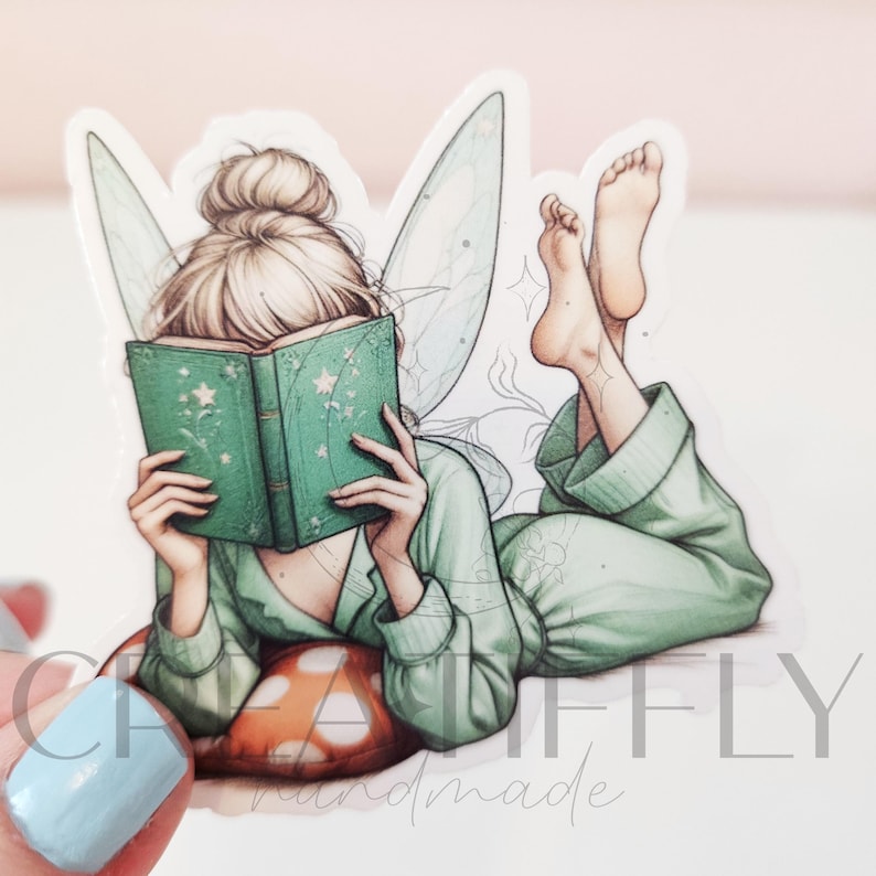 May include: A sticker featuring a fairy reading a book. The fairy is wearing a green outfit and has blonde hair in a bun. She is lying on a mushroom-shaped pillow and has her feet up in the air. The book is green with star details.