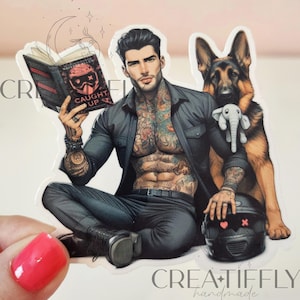 May include: A sticker featuring a tattooed person reading a book titled "CAUGHT UP." A German Shepherd dog and a black helmet with a heart and crossbones are also in the image. The sticker is labeled "CREATIFLY handmade."