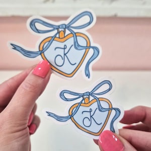 May include: Two heart-shaped stickers with a light blue background and a gold border. Each sticker has a blue ribbon and the letter "K" in the center. The stickers are held by a person with pink nail polish.