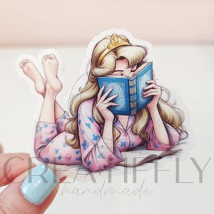 May include: A sticker featuring a cartoon illustration of a woman with long blonde hair wearing a gold crown and pink pajamas with blue butterfly patterns. She is lying down and reading a blue book.