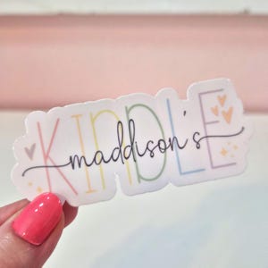 May include: A white sticker with the name "Kindle Maddison's" in a colorful, whimsical font. The word "Kindle" is in pastel colors, and "Maddison's" is in a cursive script. Small hearts and stars decorate the sticker.