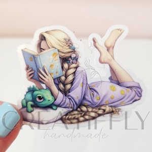 May include: A sticker featuring a cartoon illustration of a person with long blonde hair reading a book while lying down. The person wears a purple pajama set with yellow flower accents and is accompanied by a green chameleon. The sticker has the text "KREATIVFLY handmade".