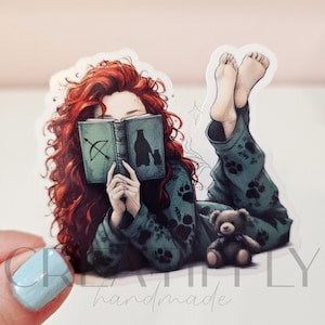 May include: A sticker featuring a person with red curly hair reading a book with an illustration of a bow and arrow and a bear. The person is wearing green pajamas with paw prints and has a small teddy bear. The text "Creatively handmade" is at the bottom.