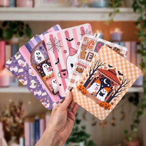May include: A set of four rectangular cards featuring illustrations of ghosts, pumpkins, and books. The designs include purple, pink, and orange color palettes with patterns like stripes, gingham, and spiderwebs, plus text reading Cozy Readers.