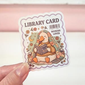 May include: A die-cut sticker shaped like a library card with a scalloped purple border. It features a white duck wearing a yellow sweater, wrapped in a colorful patchwork quilt, reading a book titled Stargazing Tales. Includes flowers, a crescent moon, a barcode, and the text LIBRARY CARD and BOOKISH LOVER.