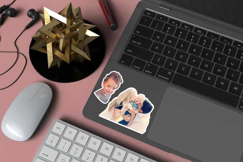 May include: A laptop computer with a sticker of a family on the lid. The sticker shows a mother, father, and two children. The laptop is on a pink surface with a white mouse, a gold geometric sculpture, and a pair of headphones.