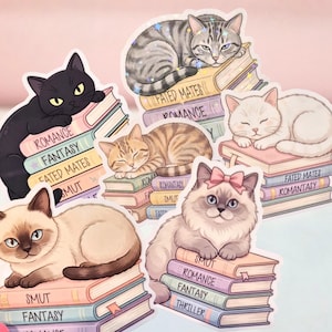 May include: A collection of six die-cut stickers featuring various cats resting on stacks of books. The books have spines labeled with genres including Romance, Fantasy, Fated Mates, Smut, Romantasy, and Thriller. The cats are depicted in different colors.