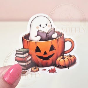 May include: A sticker featuring a cute white ghost reading a book while sitting in an orange jack-o'-lantern mug. Small pumpkins and a stack of books are next to the mug. The sticker has a Halloween theme.
